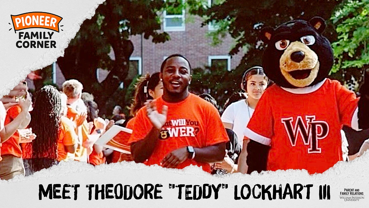 A person and a mascot are standing together at an outdoor event, both wearing orange shirts, with a crowd in the background.